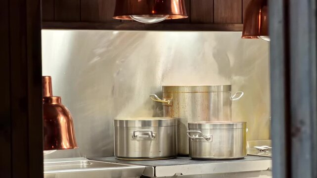 Steam rises from large metal pots on a commercial kitchen stove under copper lights