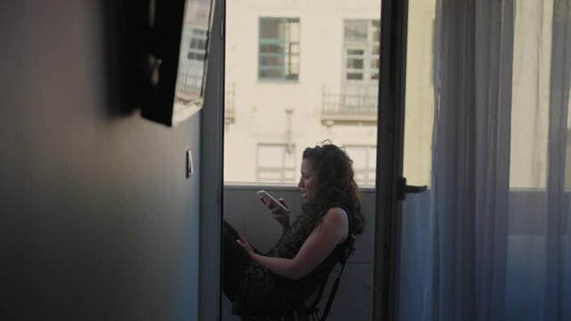Young woman sitting on small European balcony in her apartment recording audio message on mobile phone. Summer day as female tourist or local enjoying shade talking to friends or family over phone