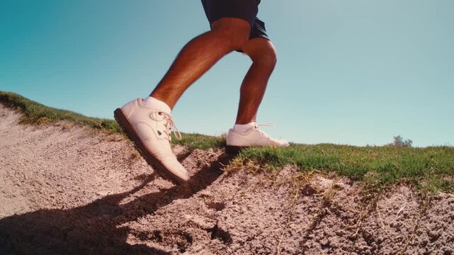 A low angle frames a golfer's white shoes stepping up out of a sand bunker with club in hand against a clear blue sky in slow motion, a gritty ground level look for sports edits.