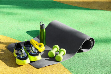 Fototapeta premium Fitness mat with sneakers, headphones, water bottle and dumbbells on sports ground