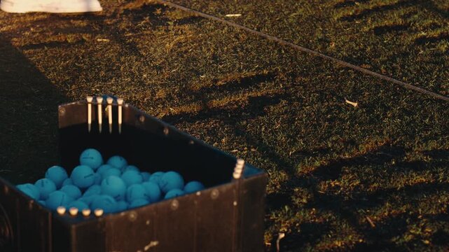 Range balls piled in a wooden basket with tees on driving range turf at golden hour in slow motion. Golf practice promotions, driving range marketing, sports equipment retail.