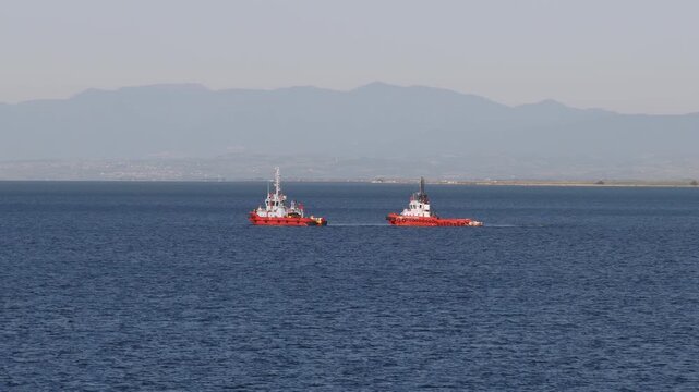 Two red tugboats sail side by side across the gulf waters near Thessaloniki, with hazy coastal mountains in the background.