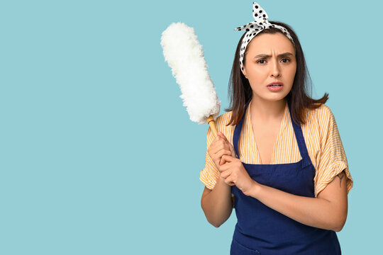 Portrait of angry young housewife in apron with pp duster on blue background