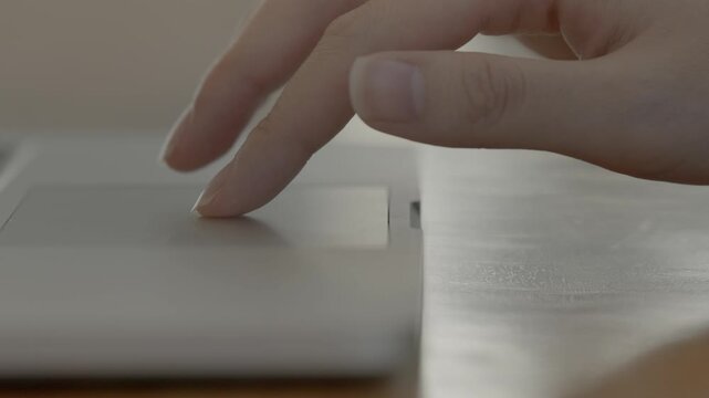 A close-up of a woman's hand using a laptop touchpad with fingers gliding over the surface in a bright workspace.