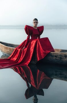 High fashion editorial portrait of a woman in an extravagant red voluminous gown sitting in a rustic wooden boat on a misty calm lake with dramatic mirror water reflection
