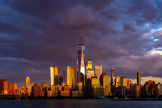 New York cityscape with storm clouds. New York golden light hits towers. New York City frames Manhattan waterfront. NYC skyline reflection on water.