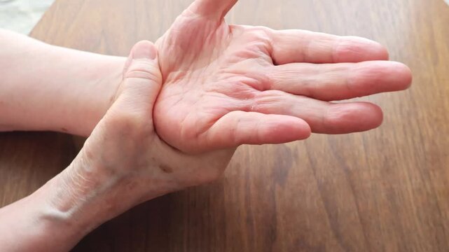 An elderly woman touching her hand on a wooden table, concerned about the pain.