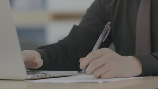 A close-up of a person holding a pen and writing notes on paper beside a laptop in a bright workspace.