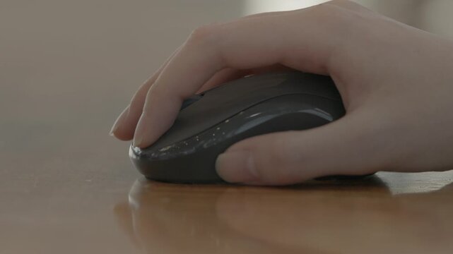 A close-up of a woman's hand gently resting on a dark wireless mouse on a desk in a bright professional workspace.