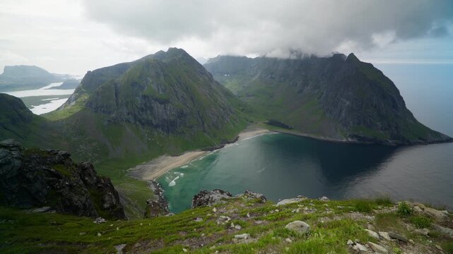 Rocky ridge foreground frames Kvalvika Beach and its curved sand bay enclosed by steep green mountain walls under a low gray sky.