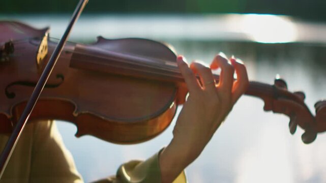 Close-up of a musician hand gracefully playing a wooden violin outdoors near water, capturing delicate strings, bow movement, and warm natural sunlight reflections