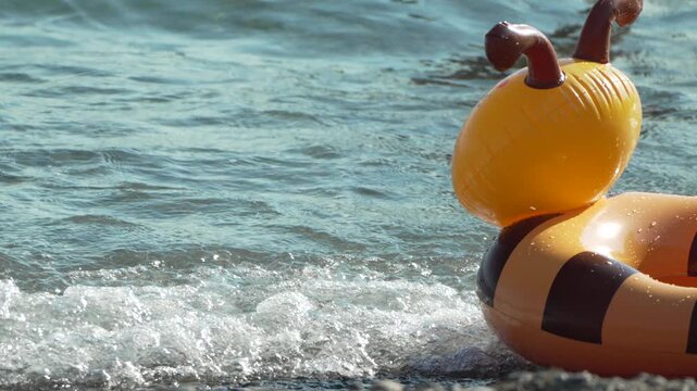 Bee shaped pool float rocks on the shore as gentle waves roll onto the pebble beach. Static eye-level shot.