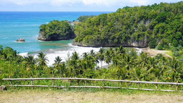 High-angle scenic view of a tropical bay with turquoise waters, dramatic limestone cliffs, and lush green palm forests on the southern coast of Pacitan, East Java, Indonesia.