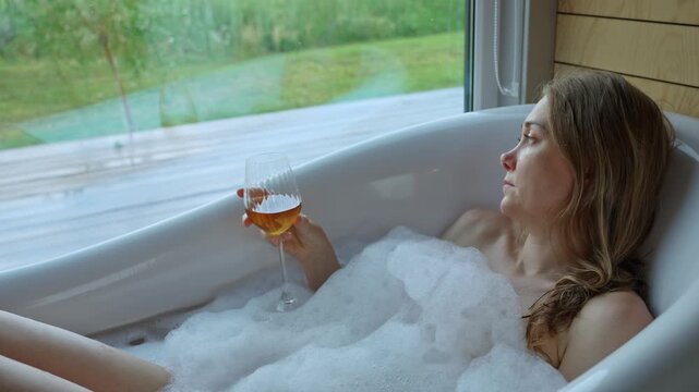 A calm woman takes a lavish bubble bath, her hand gracefully holding a wine glass. She contemplates the verdant outdoor scenery from her modern bathroom window during a quiet day.