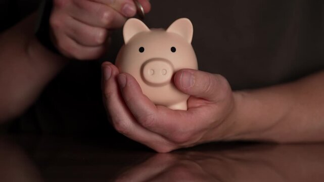 A person's hands carefully deposit a bright coin into a cute, pink piggy bank held on a polished table.