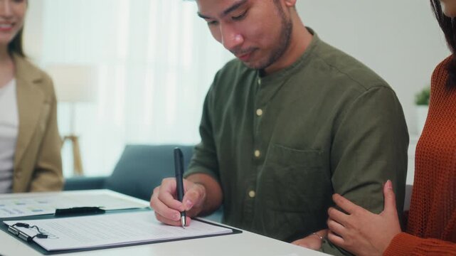 Closeup young Asian couple signing contract paper with advisor in living room at home. Financial investment consultation, house mortgage loan agreement signature, property insurance deal concept.