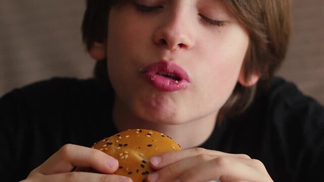 Close-up of a young boy eating a delicious burger with his eyes closed in pure delight. The child happily takes a bite of juicy fast food during a satisfying snack break.