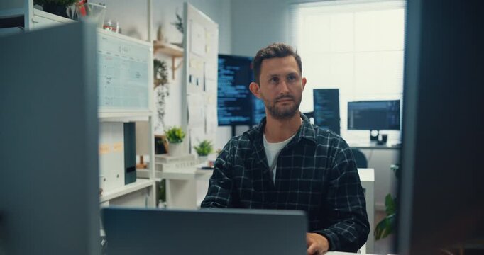 Young Caucasian male programmer writes code using multiple screens in home office workspace. AI development, cybersecurity, network database system, software engineering, remote tech startup concept.