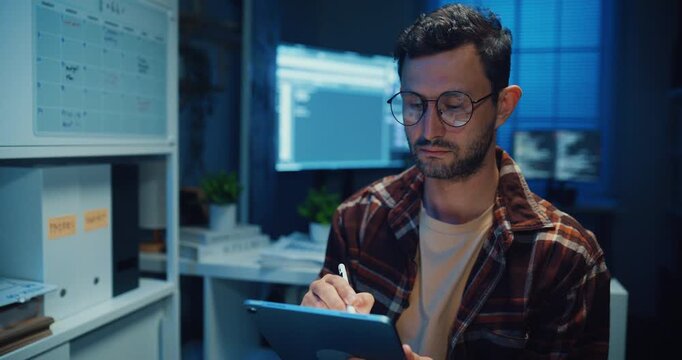 Young Caucasian male programmer use tablet for code design and plan in dark home office at night. AI technology, cybersecurity, software developer, freelance innovation, digital data network concept.