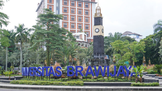 A iconic view of the Universitas Brawijaya campus entrance in Malang, Indonesia, featuring the prominent blue university signage, a decorative clock tower monument, and lush tropical landscaping.