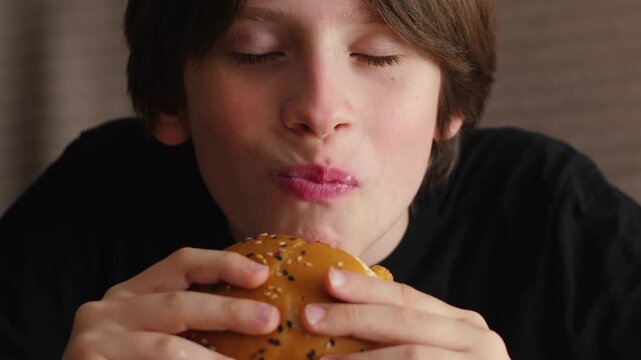 Close-up of a young boy taking a bite and chewing a juicy hamburger with sesame seeds. The kid closes his eyes with pleasure, enjoying the taste of his favorite fast food