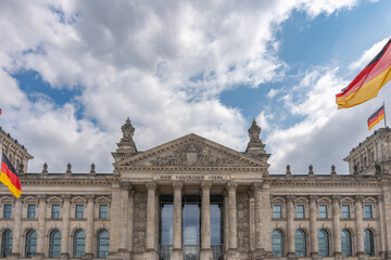 Naklejka premium The Reichstag's symmetrical front facade stands beneath dramatic clouds. German flags frame the entrance and the inscription Dem Deutschen Volke in Berlin.