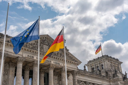 European Union and German flags wave before the Reichstag facade. Classical columns, sculpture and clouds form a strong image of European and national government.