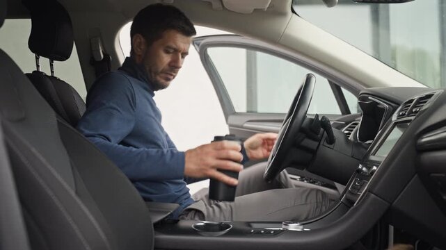 Man gets into the driver's seat with a reusable coffee cup places it in the cup holder closes the door and fastens his seat belt before holding the steering wheel.