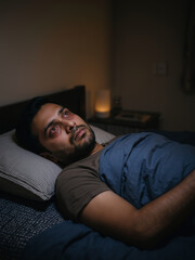 Obraz premium South asian man lying awake in bed at night with red bloodshot eyes. Sleeplessness and insomnia health issues vertical banner with copy space
