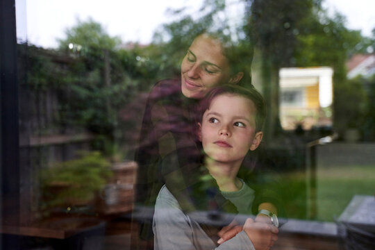 Woman with eyes closed embracing son at home at home seen through window
