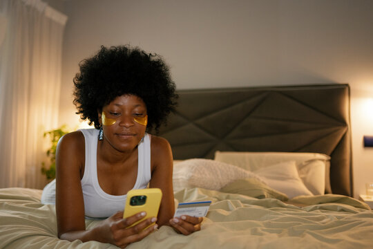Woman relaxing in bed using smartphone and credit card in morning
