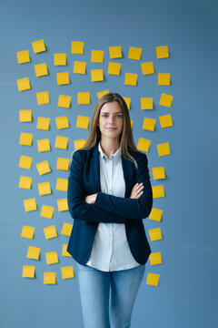 Yong businesswoman standing in front of wall, full of yellow sticky notes, with arms crossed