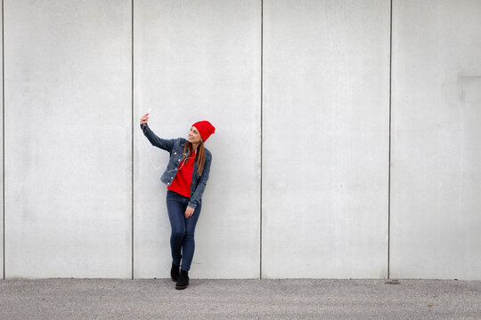 Woman wearing red pullover and wolly hat and taking a selfie in front of a wall