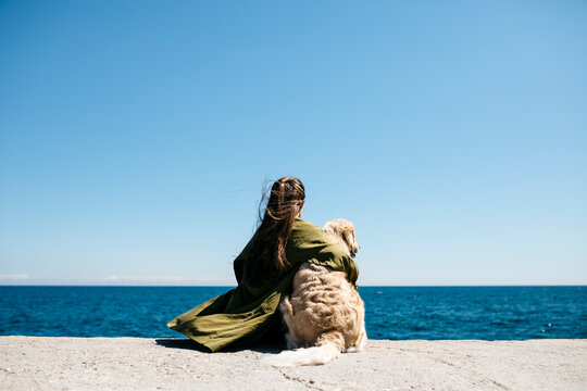 Back view of woman sitting besides her Labrador Retriever at quay looking at horizon