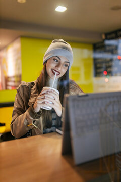 Woman holding drink looking at laptop in cafe seen through glass