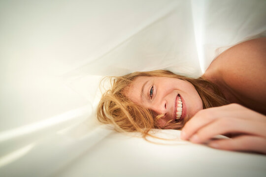 Portrait of happy blond young woman lying in bed under blanket