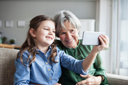 Grandmother and her granddaughter sitting together on the couch taking selfie with smartphone