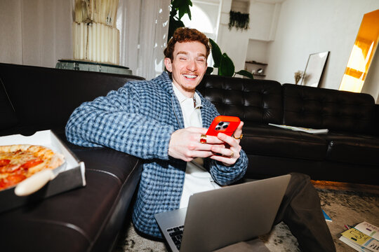 The man balances smartphone use with laptop work on the living room floor at home.