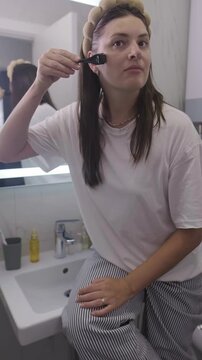 Young woman in cozy pajamas performs a home beauty routine in the bathroom, using a microneedle roller on her face as part of her skincare ritual and self care routine. 