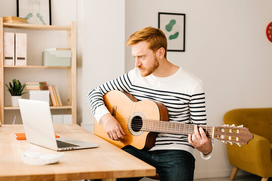 Young man with guitar e-learning through laptop sitting at table