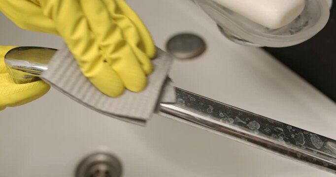 Cleaning the bathroom faucet, hands in yellow gloves wipe the faucet with a cleaning solution.