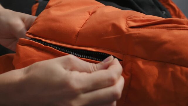 Woman operating zipper on orange jacket with focus on fastening mechanism. Zipper showcases reliable performance, quality hardware, durable construction, quilted outerwear and modern product. 4K video