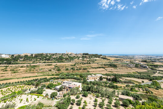 Landscape views of central Malta from the fortified walls of Mdina, showing rolling farmland, scattered houses, green valleys and a distant coastline under bright Mediterranean sunlight.