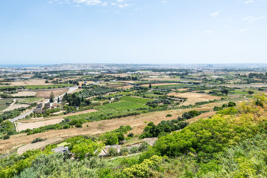 Panoramic landscape views of central Malta from the fortified walls of Mdina, showing rolling farmland, scattered houses, green valleys and a distant coastline under bright Mediterranean sunlight.