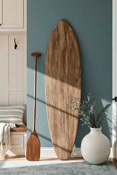Weathered wooden paddle board and oar leaning against a matte teal wall in a modern coastal mudroom with soft morning light