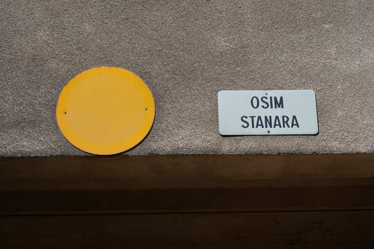 A close up shot of a completely faded yellow round no entry traffic sign next to a white plate with Croatian text OSIM STANARA meaning except residents on a grey wall