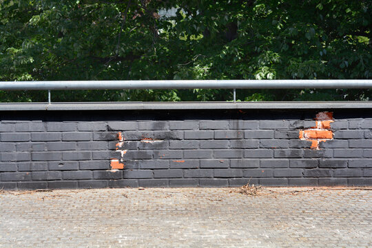 A horizontal view of a black painted brick parapet wall with damaged sections revealing orange bricks below a metal railing and green trees