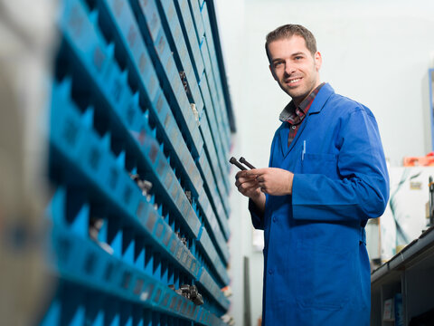 Portrait of a smiling male worker in a blue uniform holding small metal bolts or screws in a workshop or hardware store warehouse, standing next to structured storage shelves.