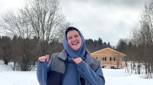 Smiling youth in blue winter jacket raises fist outdoors in snowy yard under overcast sky, with leafless trees and rustic cabin creating cheerful seasonal atmosphere