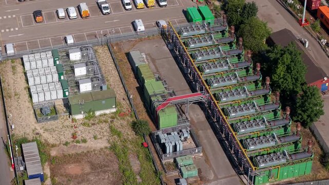 Aerial view tracking over a complex arrangement of commercial power generation machinery enclosures, exhaust systems, and service lines inside a rail maintenance yard.